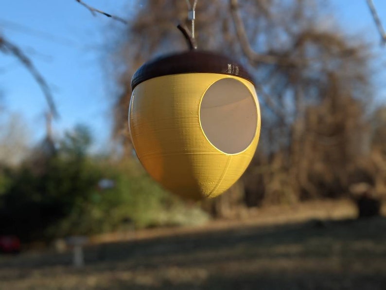 May include: A yellow and brown acorn-shaped birdhouse hanging from a tree branch. The birdhouse has a circular opening and a textured surface. The top is brown, and the body is yellow. The background is blurred, showing a blue sky and greenery.