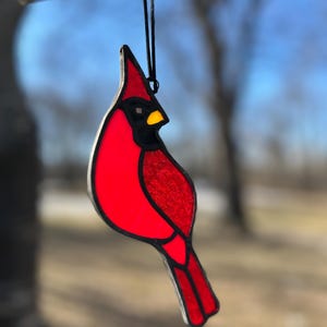 Op de afbeelding: Een ornament van gekleurd glas in de vorm van een kardinaal met een rood lichaam, een zwarte omtrek en een gele snavel. Het ornament hangt aan een zwart touwtje.