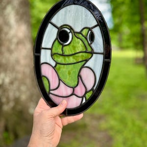 May include: Oval stained glass art piece featuring a green frog with large eyes and a pink dress. The frog is set against a white and clear background, framed by a black border. A whimsical decorative item.