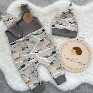 May include: A gray and white baby onesie with a construction vehicle print. The onesie has a bib attached and a gray hat with the same print. A wooden sign with the text "Liam & Lippy" is in the background.