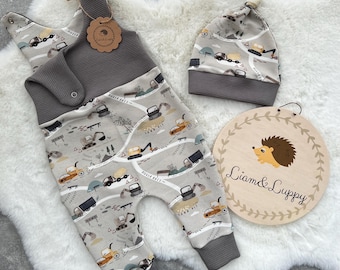 Op de afbeelding: Een grijze en witte baby romper met een print van bouwvoertuigen. De romper heeft een bijgevoegde slabbetje en een grijze muts met dezelfde print. Een houten bord met de tekst "Liam & Lippy" is op de achtergrond.