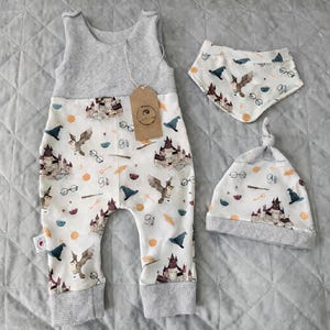 Puede incluir: Un conjunto de ropa de bebé con un pelele, un babero y un gorro. La tela blanca está estampada con un castillo, un búho, gafas y otros elementos mágicos. El pelele tiene una parte superior de punto de gofre gris.