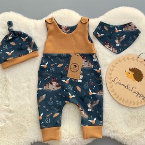 May include: A baby outfit set featuring a dark teal romper, hat, and bib with a repeating pattern of castles, owls, and potion bottles. The set includes a mustard yellow waffle-knit top and trim. A wooden sign with a hedgehog illustration and the text "Liam & Luppy" is also included.