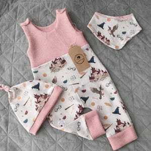 Puede incluir: Un conjunto de ropa de bebé con una parte superior de punto de gofre rosa y pantalones estampados, además de un babero y un gorro a juego. El estampado incluye un castillo, búhos, gafas y elementos mágicos. El conjunto está diseñado para bebés.