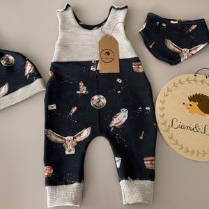 Könnte beinhalten: Ein Baby-Kleidungsset mit einem marineblauen Strampler, Hut und Lätzchen mit einem skurrilen Eulen- und Himmelsdruck. Der Strampler und der Hut haben einen Waffelstrickbesatz. Ein Holzschild mit einer Igel-Illustration ist ebenfalls enthalten.