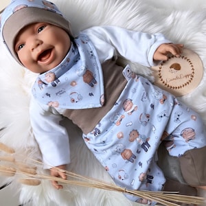 May include: A baby doll wearing a blue and brown outfit with a cute animal print. The outfit includes a hat, bib, and pants. The doll is lying on a white fluffy surface.