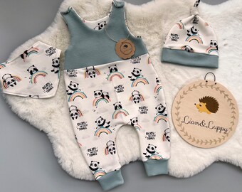 Op de afbeelding: Een babykleding set met een panda print in lichtblauw en wit. De set bevat een jumpsuit, slab en muts, allemaal versierd met panda's, regenbogen en de tekst "Enjoy the little things". Een houten bord met een egel illustratie is ook aanwezig.