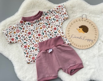 May include: A baby's two-piece outfit set. The top is white with a floral pattern in pink, brown, and green. The shorts are mauve. A wooden sign with the text "Liam & Luppy" and a hedgehog illustration is also in the image.