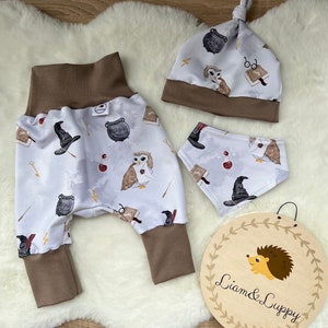 May include: A baby outfit set with a white and brown owl, book, and cauldron print. The set includes a hat, pants, and a bib. The hat has a brown brim and a white top with a brown owl, book, and cauldron print. The pants are brown with a white and brown owl, book, and cauldron print. The bib is white with a brown owl, book, and cauldron print. The set is made of soft, comfortable fabric and is perfect for a baby boy or girl.
