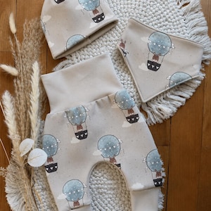 Può includere: Un completo di abbigliamento per neonati beige con un motivo a mongolfiera. Il set include un berretto, pantaloni e un bavaglino. Le mongolfiere sono azzurre con un animale marrone nel cestino. I pantaloni hanno una vita alta.