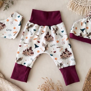 May include: A baby clothing set comprising trousers, a bib, and a hat, all with a white background and a repeating pattern of a castle, owls, and other magical elements. The set has burgundy accents.