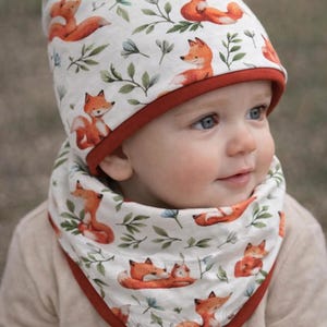 May include: A baby wearing a white hat and bib set. The hat and bib feature a repeating pattern of orange foxes and green leaves on a white background. Both items have a red trim.