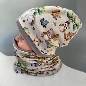 May include: A beige knit hat and matching neck warmer with a woodland animal print. The print features a variety of animals including owls, deer, raccoons, and bears. The hat and neck warmer are both trimmed with a gray band.