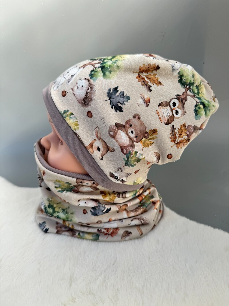 May include: A beige knit hat and matching neck warmer with a woodland animal print. The print features a variety of animals including owls, deer, raccoons, and bears. The hat and neck warmer are both trimmed with a gray band.