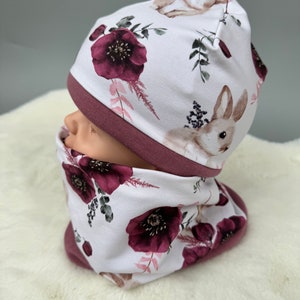 Könnte beinhalten: Eine weiße Mütze und ein passender Halsschmuck mit einem Blumenmuster mit burgunderfarbenen Blumen, grünen Blättern und weißen Hasen.