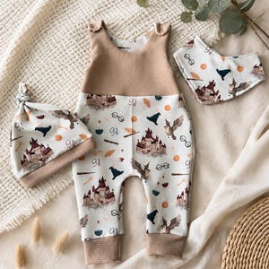Könnte beinhalten: Ein Baby-Strampler, eine Knotenmütze und ein dreieckiges Lätzchen mit einem Muster aus Schlössern, Eulen, Brillen, Zauberstäben und dem Text 9 3/4. Der Strampler hat ein beiges Waffelstrick-Oberteil und Bündchen, die anderen Teile sind weiß.