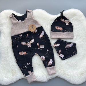 May include: A baby outfit set featuring a black jumpsuit, a matching beanie, and a bib. The set is adorned with a whimsical print of owls, letters, and celestial elements. The jumpsuit and beanie have a beige trim.