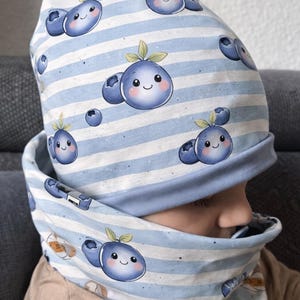 May include: A blue and white striped hat and neck warmer set. The fabric is patterned with smiling cartoon blueberries. The hat is a beanie style, and the neck warmer is a circular scarf. The set is made of soft fabric.