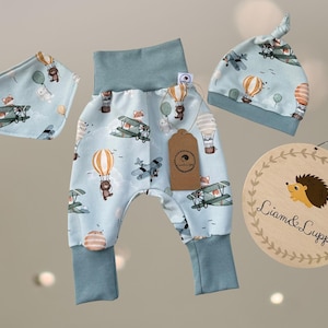 Può includere: Un set di abbigliamento per bambini in azzurro con un motivo di mongolfiere, aerei e animali. Il set comprende pantaloni, un bavaglino e un cappello annodato. È presente anche un cartello in legno con un'illustrazione di riccio.