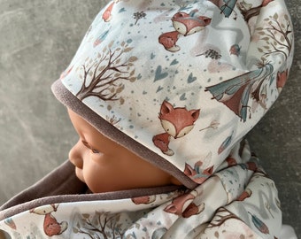 May include: A white baby hat and neck warmer set with a fox and teepee print. The fabric is soft and comfortable.