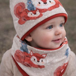 May include: A beige baby hat and neck warmer set with a repeating pattern of red foxes and blue birds. The hat and neck warmer have a red trim. The fox illustrations have white faces and are lying down. The background has small white polka dots.