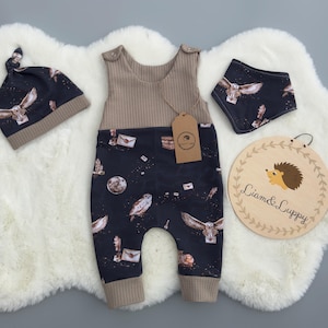 May include: A baby outfit set featuring a brown waffle knit overall with a black and white owl print, a matching black and white owl print hat, and a black and white owl print bib. The set is laid out on a white fluffy surface.