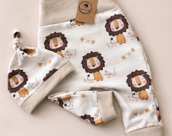 Op de afbeelding: Een baby-outfitset met een leeuwenprint. De set bevat een mouwloze romper en een geknoopte muts. De romper en muts zijn wit met een streepjespatroon en leeuwenillustraties. De afwerking is lichtbruin.