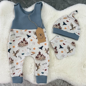 May include: A baby outfit set featuring a blue waffle-knit top and patterned pants with a matching hat and bib. The pattern includes a castle, owls, glasses, and other magical elements. A brown tag hangs from the outfit.