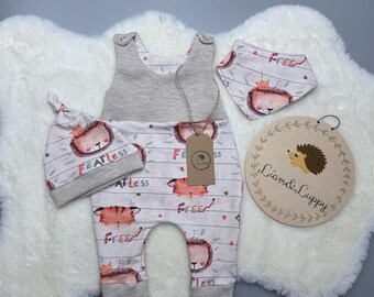 Op de afbeelding: Een baby-outfitset met een grijs-wit gestreepte romper met een leeuwenprint, een bijpassende muts met de tekst "FEATLess" en een slab met een leeuwenprint. De set ligt op een wit, pluche oppervlak.