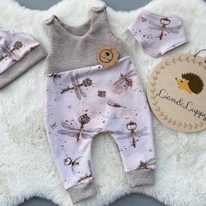 May include: A baby outfit set with a beige and pink jumpsuit, a beige hat with a dragonfly design, and a pink and white bib with a dragonfly design. The jumpsuit has a dragonfly and key design. The set is on a white fluffy surface.