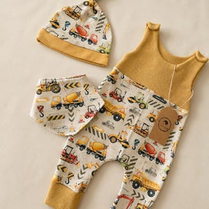 Puede incluir: Un conjunto de ropa de bebé con gorro, babero y mono. El conjunto tiene un estampado de vehículos de construcción sobre un fondo crema, con detalles en amarillo mostaza. El mono tiene una parte superior de punto de gofre. El conjunto es para bebés.