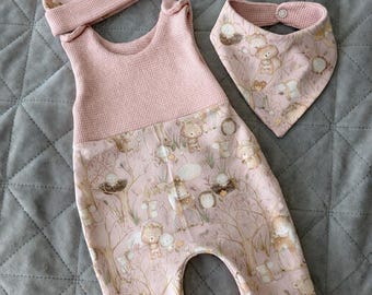 May include: A baby clothing set in pink, featuring a hat, jumpsuit, and bib. The jumpsuit and bib have a woodland animal print. The hat and jumpsuit have a waffle knit texture. The set is designed for infants.