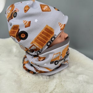 May include: Gray and brown beanie with a construction truck pattern. The beanie has a matching neck warmer with the same pattern.