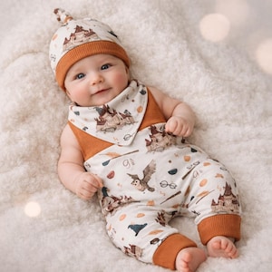 May include: A baby wearing a matching set of clothing, including a hat, bib, and jumpsuit. The outfit is white with a brown castle and owl pattern, and brown trim. The baby is lying on a white, fluffy surface.