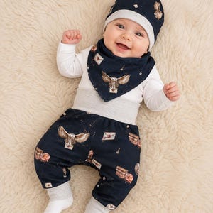 Könnte beinhalten: Ein Baby in einem marineblauen Hut, Lätzchen und Hosen-Set mit einem sich wiederholenden Muster aus Eulen, Briefen und Paketen. Das Baby trägt außerdem ein weißes Langarmshirt und graue Socken. Das Baby liegt auf einer weißen, flauschigen Oberfläche.