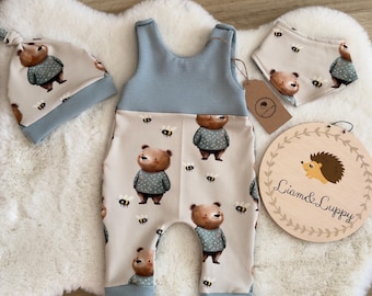 Op de afbeelding: Een baby-outfitset met een blauwe en beige romper, een bijpassende muts en een slab. De romper heeft een beer- en bijenprint. De muts en slab hebben een beerprint. De set is gelabeld met "Liam & Lucy".