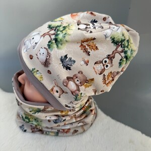 May include: A beige knit hat and matching neck warmer with a woodland animal print. The print features a variety of animals including deer, owls, raccoons, squirrels, and bears. The hat and neck warmer are both trimmed with a gray band.