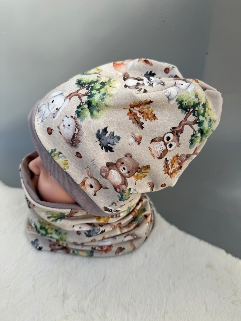May include: A beige knit hat and matching neck warmer with a woodland animal print. The print features a variety of animals including deer, owls, raccoons, squirrels, and bears. The hat and neck warmer are both trimmed with a gray band.