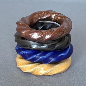 May include: A stack of four circular rings with a twisted, rope-like texture. The top ring is a metallic brown, the second is a glossy black, the third is a vibrant royal blue, and the bottom ring is a pale, speckled cream color.