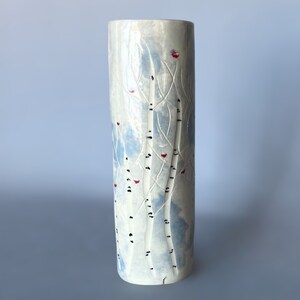 May include: White ceramic vase with a blue and white birch tree design and red accents. The vase is cylindrical in shape.