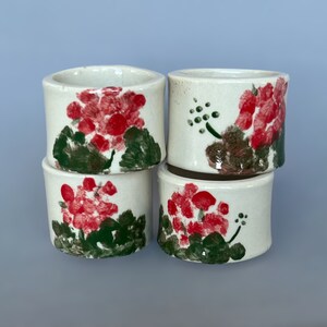 May include: Four small, white ceramic cups stacked together. Each cup is hand-painted with a red floral design, green leaves, and brown stems. The cups have a cylindrical shape and a slightly rough texture, suggesting a handmade quality.
