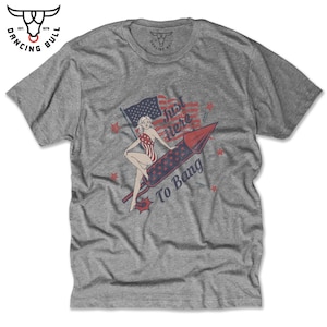 May include: A grey t-shirt with a vintage-style illustration of a woman sitting on a firework rocket. The rocket is red and white with stars and the text "Just Here To Bang".
