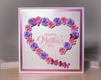 May include: A white card with a pink border and a silver background. The card has a heart shape made of pink and purple paper flowers. The text "Happy Mother's Day" is written in pink letters inside the heart.