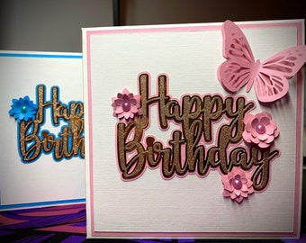 May include: Two handmade greeting cards with a pink and blue color scheme. The cards feature the words "Happy Birthday" in gold glitter lettering with decorative flowers and a butterfly.