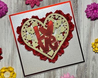 May include: A white card with a red border features a red and gold heart-shaped design with the word "Love" in red glitter letters. The heart is surrounded by red paper flowers.