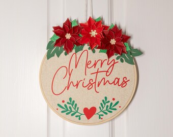 May include: A round, beige fabric wreath with the words "Merry Christmas" in red cursive lettering. The wreath is decorated with red poinsettia flowers and green leaves.
