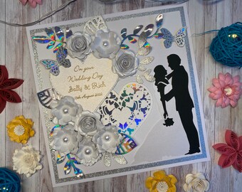 May include: A white card with a silver glitter border and a white heart with a lace design. The heart has a gold inscription that reads "On your Wedding Day Sally & Rich 13th August 2022". The card is decorated with white paper flowers and a silhouette of a bride and groom.