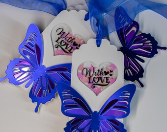 May include: Two white gift tags with blue butterfly cutouts and a heart-shaped window with pink and white confetti and the text "With Love". The tags are tied together with a blue ribbon.