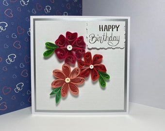 May include: A handmade birthday card featuring three quilled paper flowers in shades of red and coral, with green leaves. The card has a white background with a silver border and the words "HAPPY Birthday" in black script.