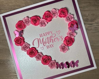 May include: A white card with a pink border and a heart made of pink and red paper flowers. The text "Happy Mother's Day" is written in pink cursive lettering inside the heart.